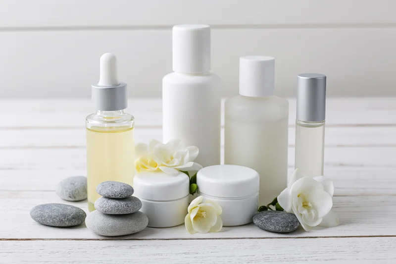 White minimalist skincare bottles and jars arranged with flowers and pebbles on a wooden surface