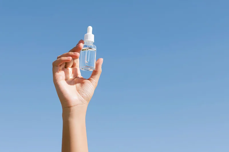 Hand holding a clear glass dropper bottle against a blue sky