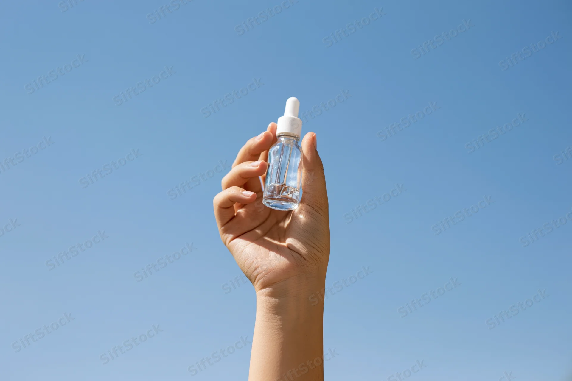 Hand holding a clear glass dropper bottle against a blue sky