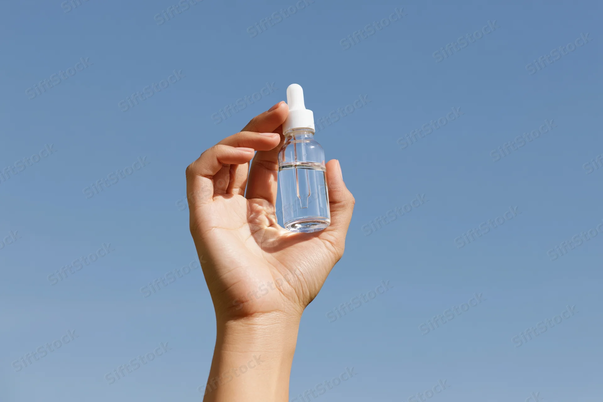 Hand holding a clear glass dropper bottle against a blue sky outdoors