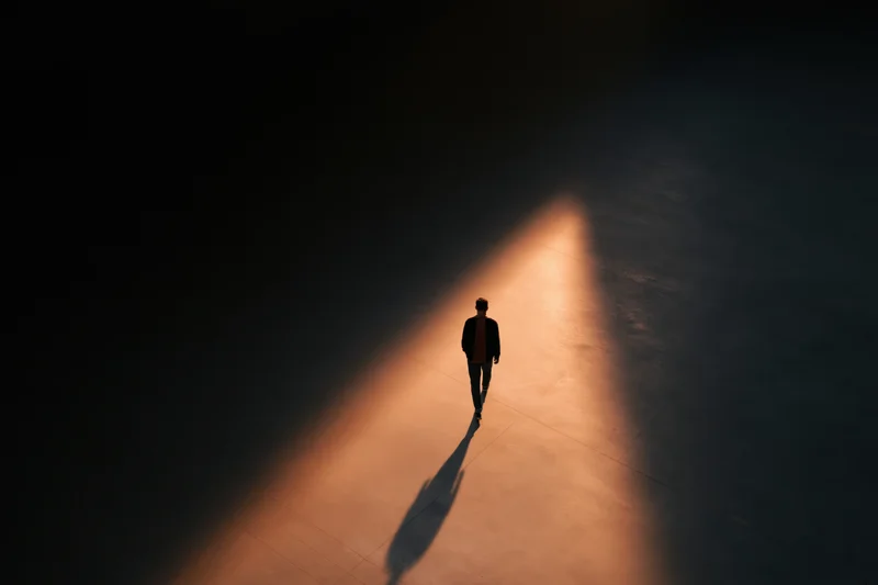 Silhouette of a man walking forward on a dark concrete path illuminated by a single beam of light