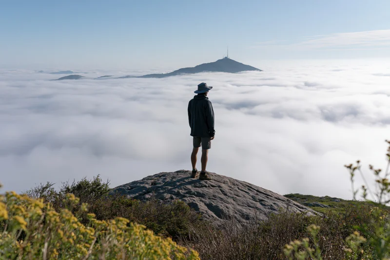 Male hiker standing on granite boulder overlooking sea of clouds from mountain summit