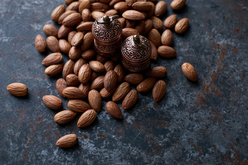 Whole unshelled almonds scattered on a dark textured surface with decorative spice jars in the center