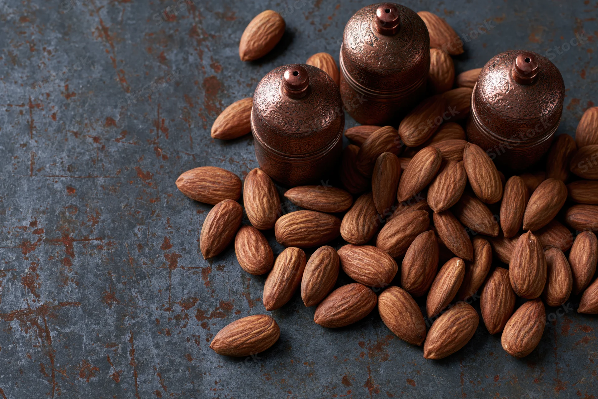 Whole unshelled almonds scattered around copper jars on a dark textured surface