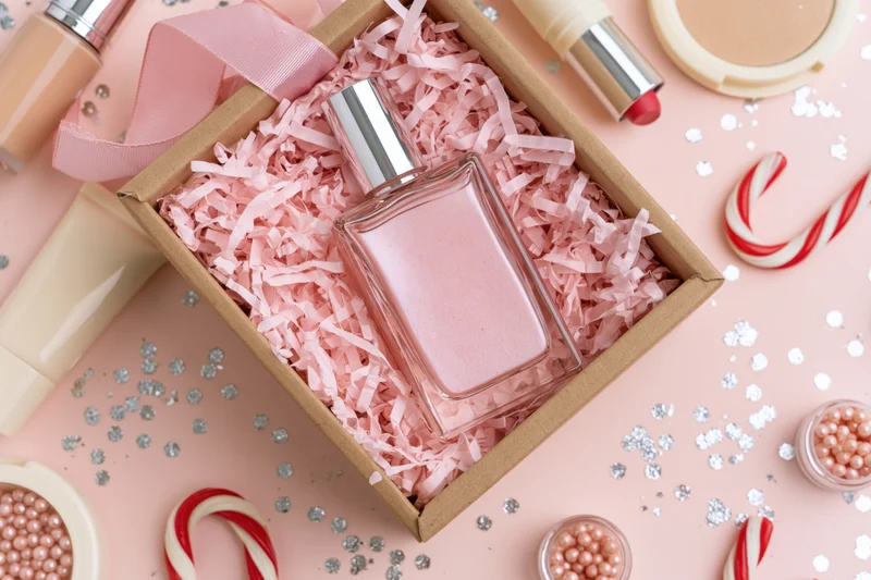 Pink glass perfume bottle in kraft gift box with shredded paper, surrounded by cosmetics and candy canes on a pink surface
