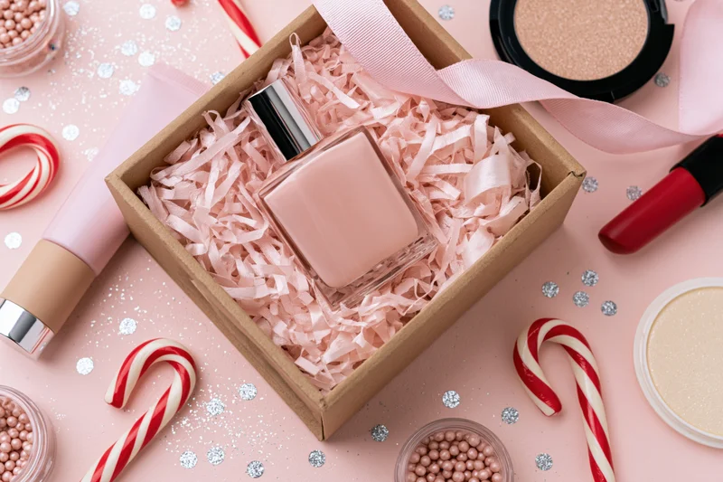 Pink perfume bottle in kraft gift box with ribbon and makeup on pink surface surrounded by candy canes and glitter