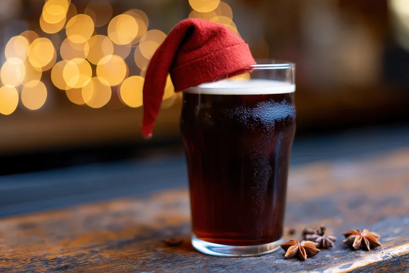 Dark amber beer in a pint glass with a small red santa hat on top, on a wooden table with star anise and blurred warm lights behind