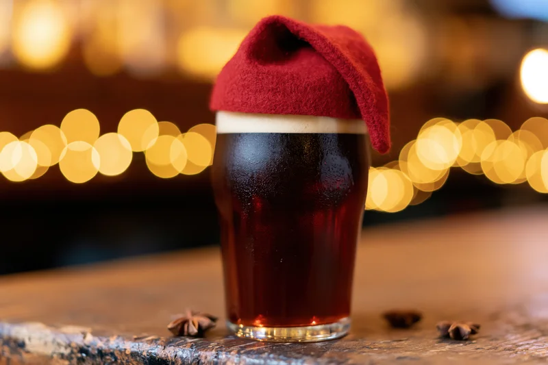 Dark amber beer in a pint glass with a red santa hat on top, on a wooden table with bokeh lights behind