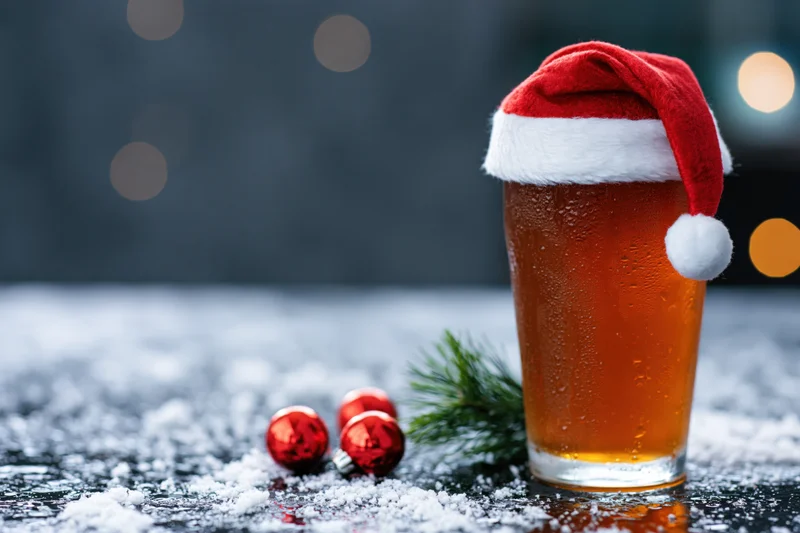 Tall glass of amber beer wearing a miniature santa hat on a snowy surface with christmas ornaments and pine sprig nearby