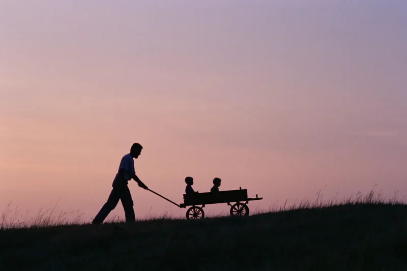 Father pulling two children in a wooden wagon across a grassy hill at sunset