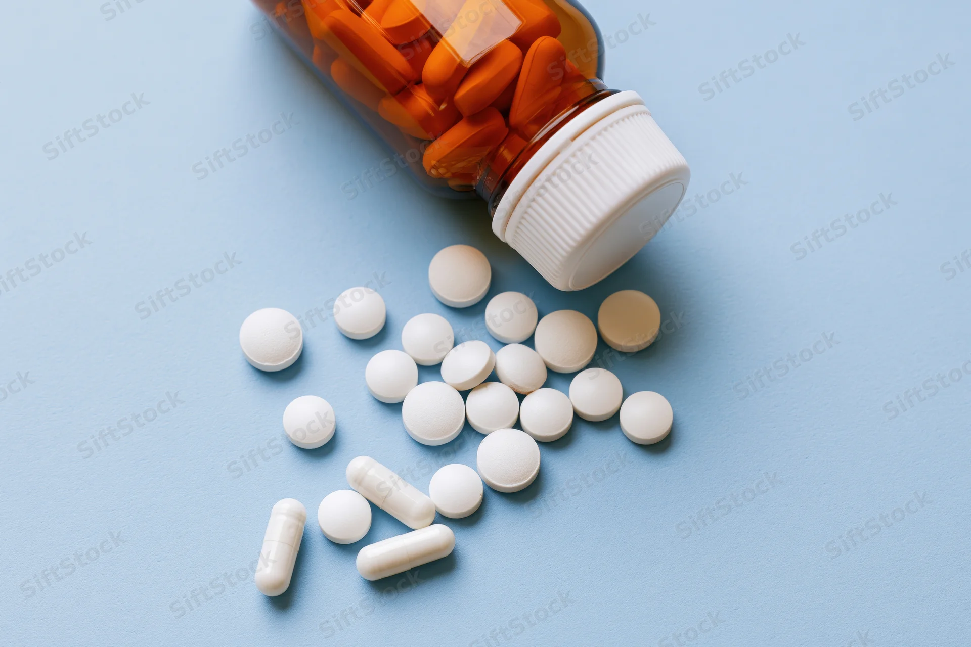 Scattered white pills and capsules from an amber glass bottle on a blue surface