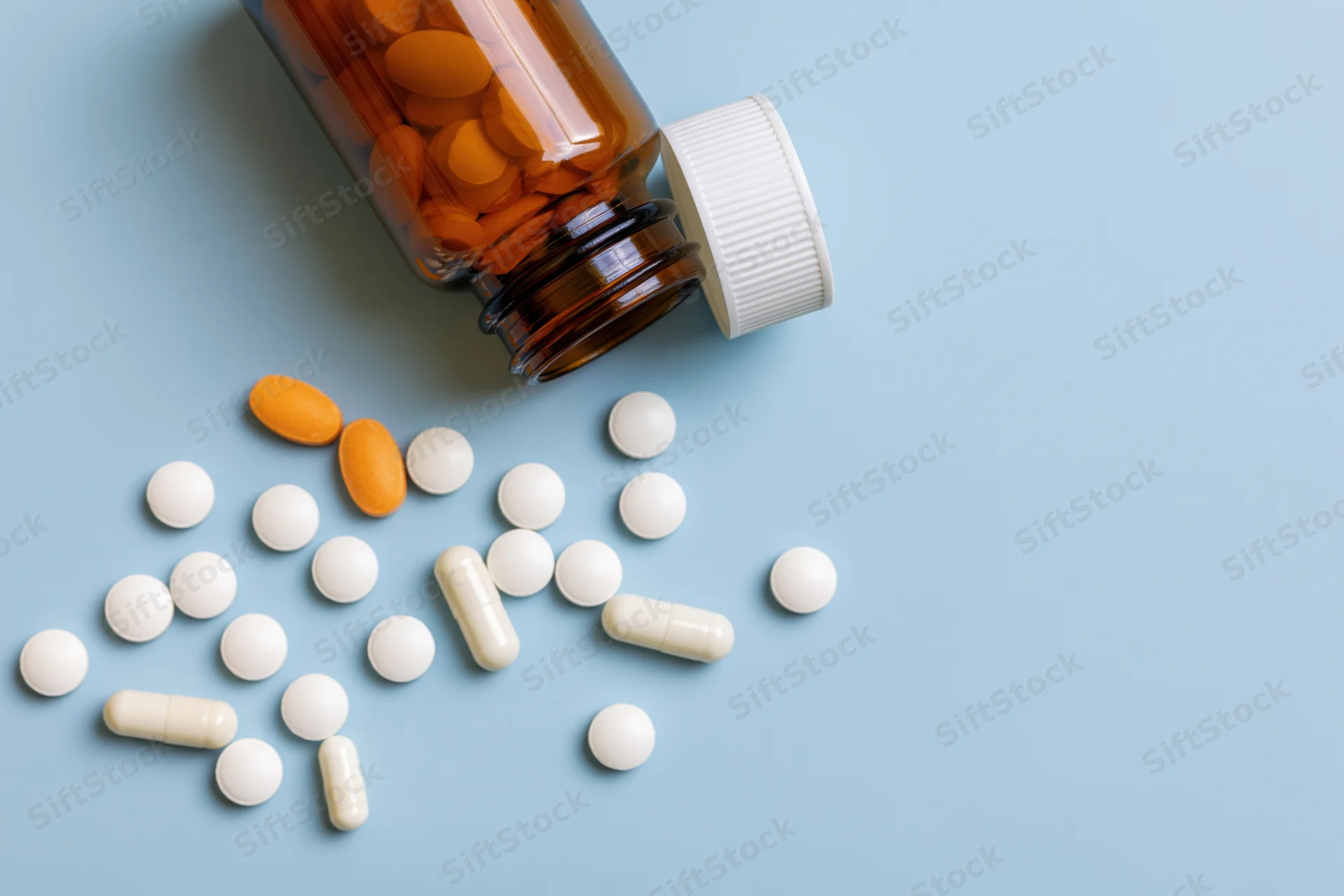 Scattered white and orange pills from an amber glass bottle on a blue surface