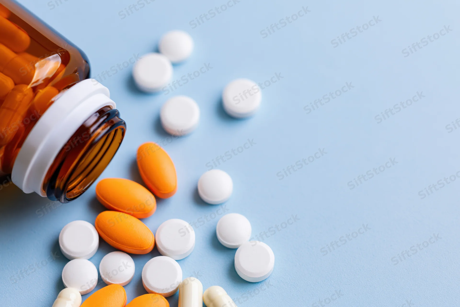 Scattered orange and white pills spilling from an amber glass bottle on a blue surface