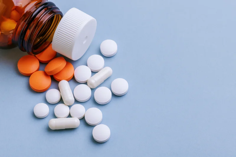 Scattered orange and white pills and capsules spilling from an amber glass bottle on a blue surface