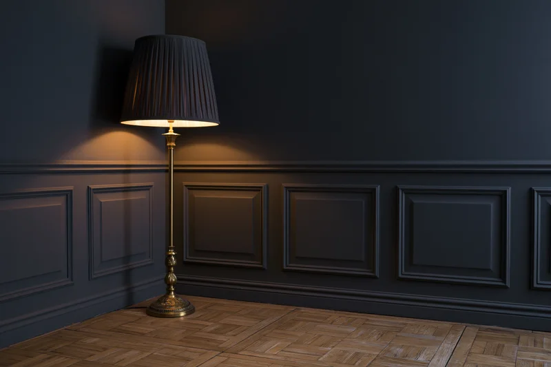 Ornate brass floor lamp with dark pleated shade glowing in a corner of a dark paneled room with parquet flooring