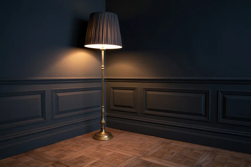 Ornate brass floor lamp with dark pleated shade glowing in a dark corner of a room with wainscoting and parquet flooring