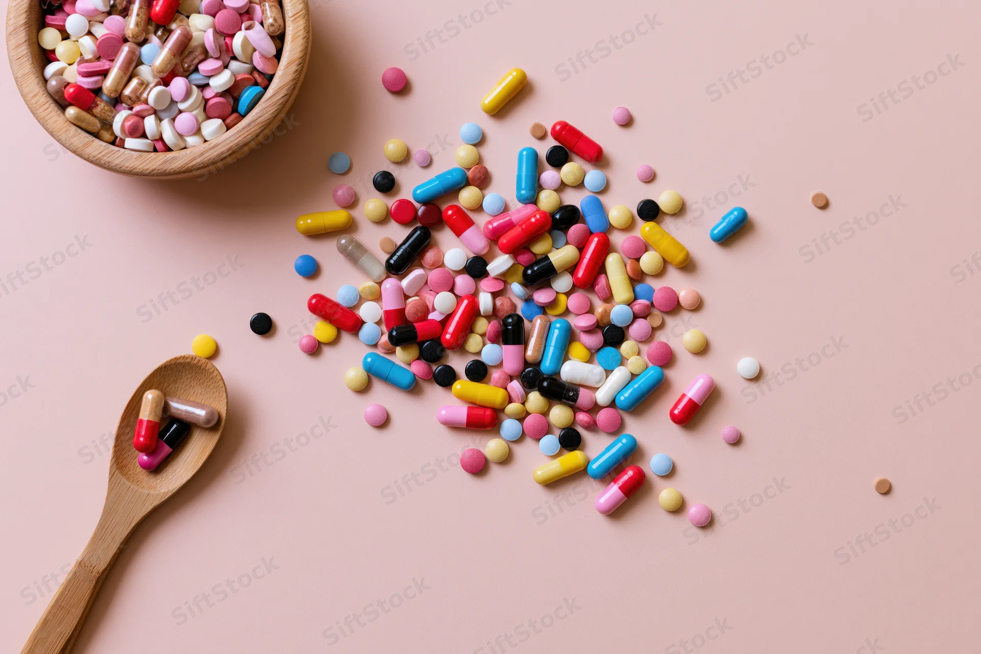 Colorful pills and capsules scattered on a pink surface with a wooden bowl and spoon