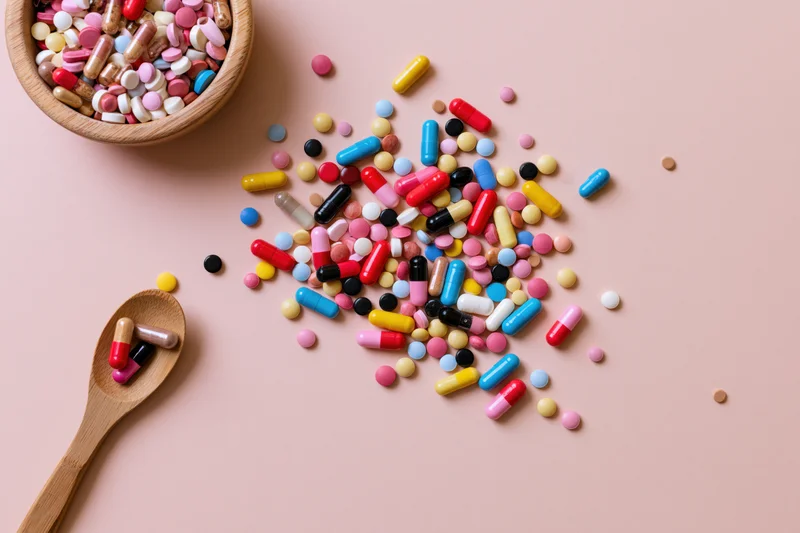 Colorful pills and capsules scattered on a pink surface with a wooden bowl and spoon