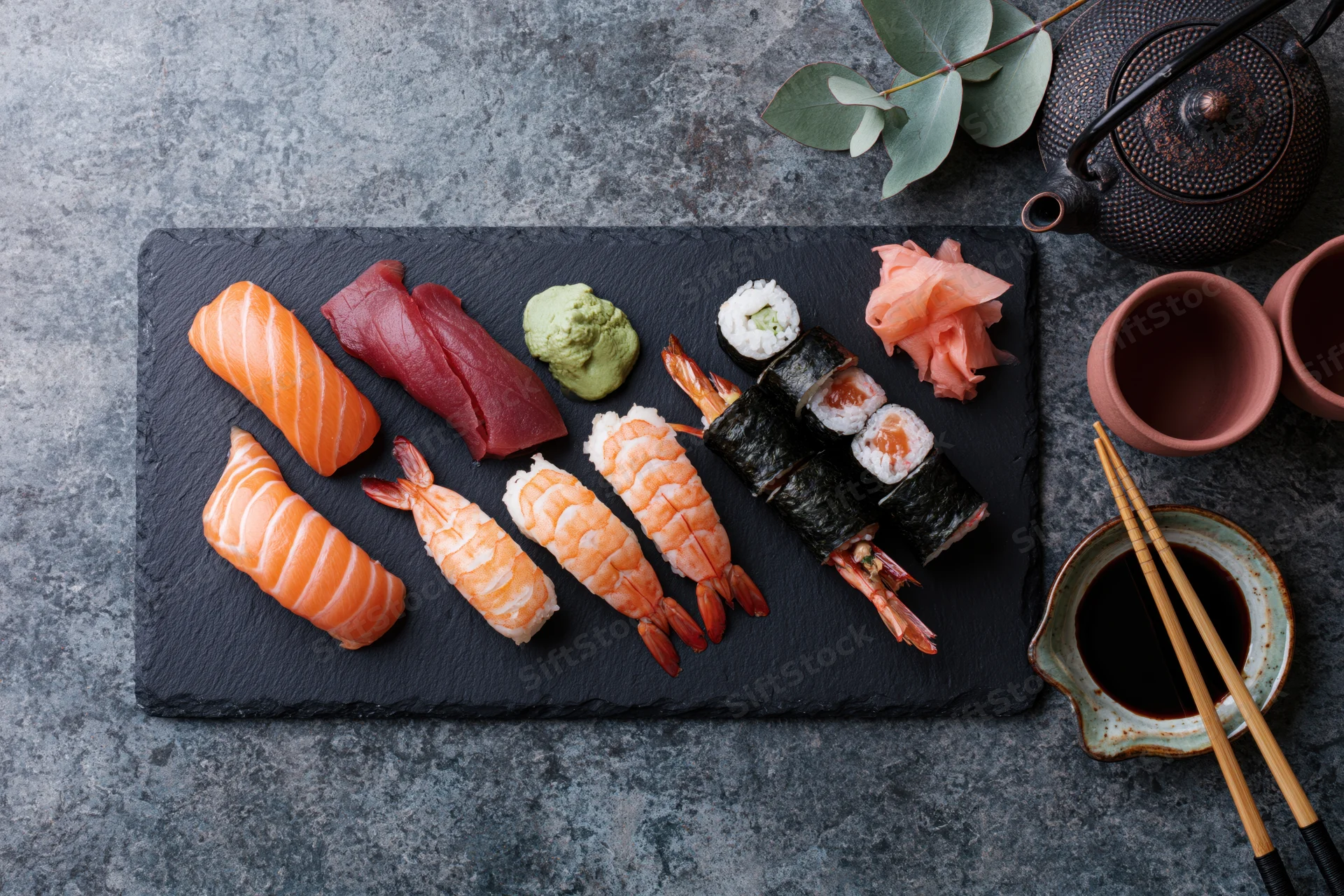 Assorted sushi arranged on a black slate platter with soy sauce, chopsticks and garnishes on a gray stone surface