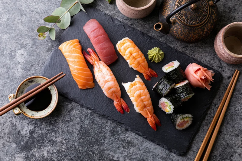 Assorted sushi pieces arranged on a black slate platter with soy sauce and chopsticks on a gray stone surface