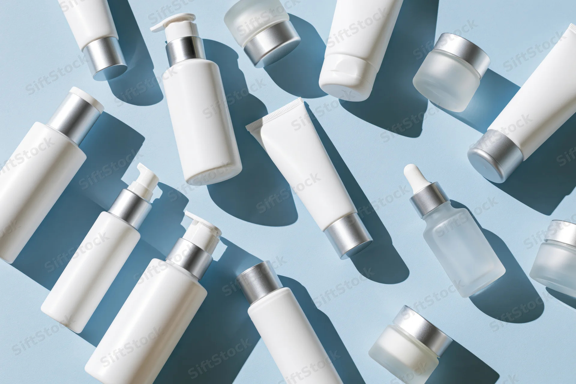 Assorted white skincare bottles tubes and jars with silver caps arranged on light blue background