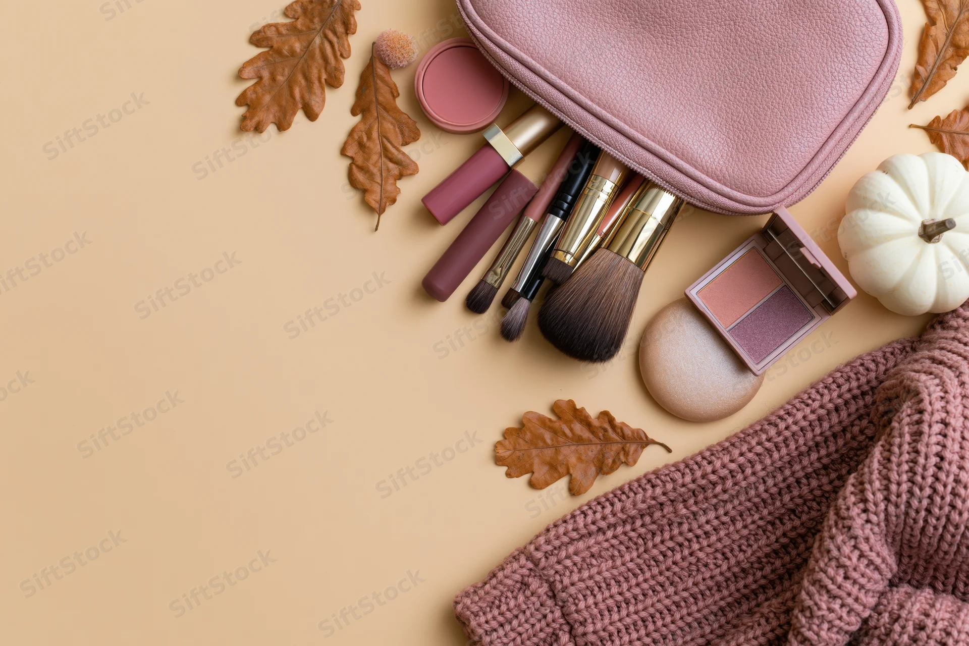 Autumn makeup flat lay with brushes, blush, lipstick and leaves on beige surface beside knitted sweater and white pumpkin