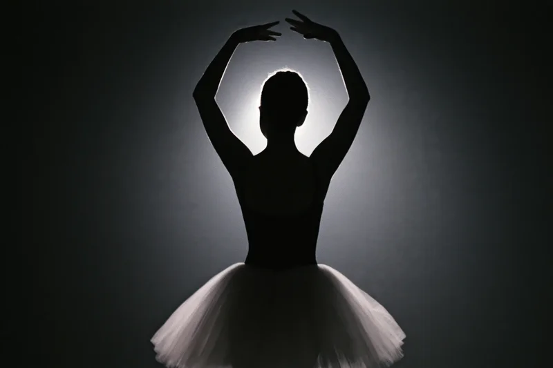 Ballerina in silhouette from behind with arms raised overhead, wearing a white tutu against a dark background