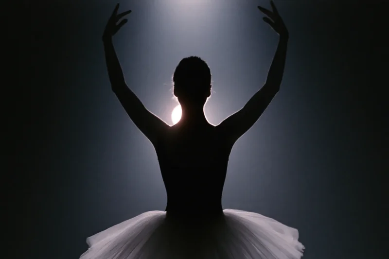 Ballerina silhouette from behind with arms raised overhead in a spotlight on stage