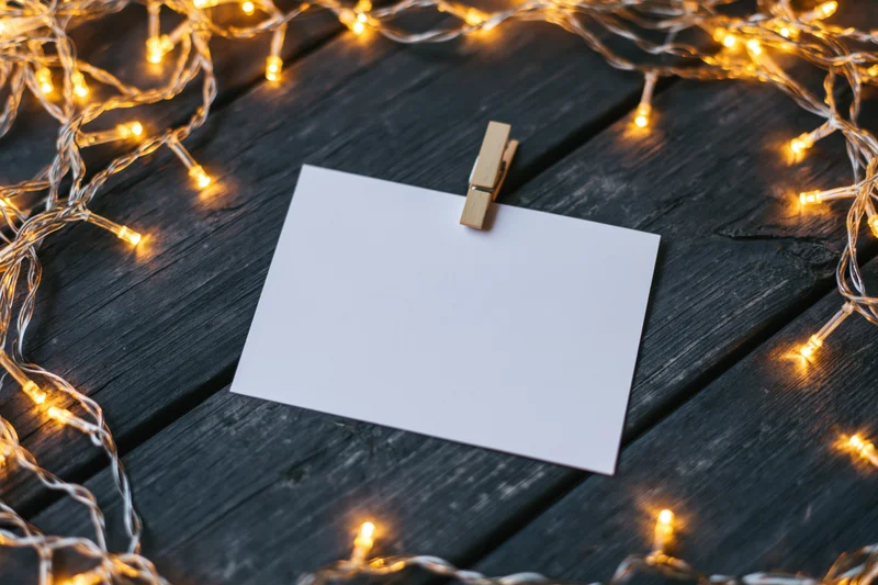 Blank white card clipped with wooden clothespin on dark wood table surrounded by warm string lights