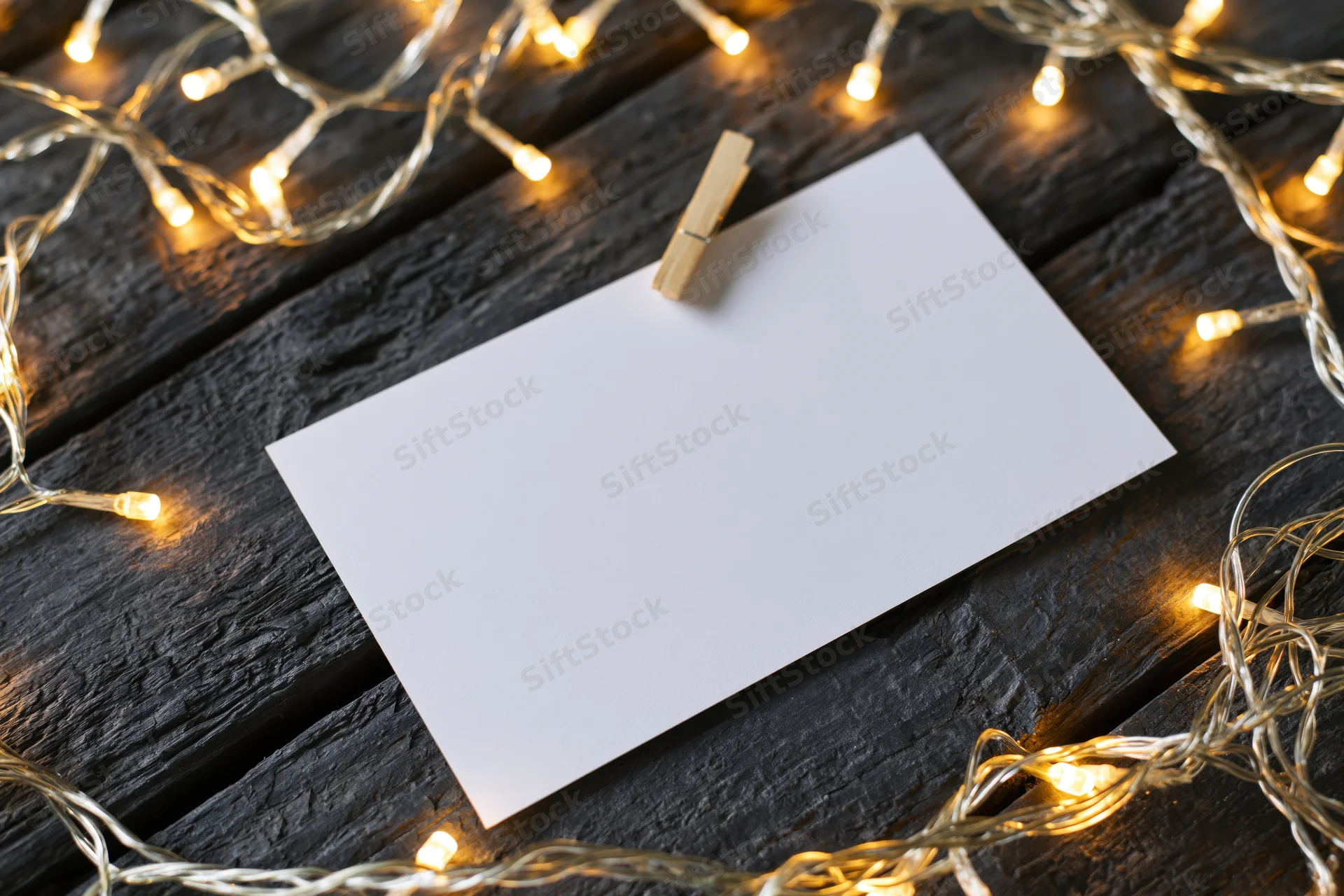 Blank white card clipped with wooden clothespin on dark wood surface surrounded by warm string lights
