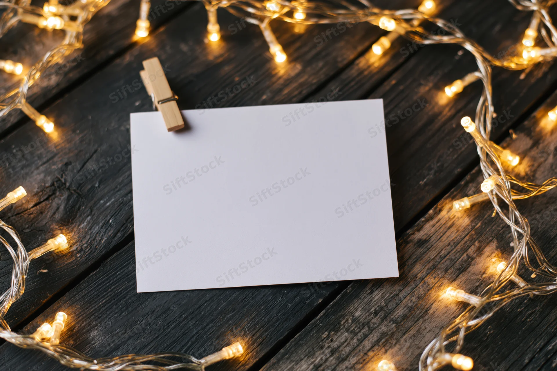 Blank white card clipped with wooden clothespin on dark wood surface surrounded by warm string lights