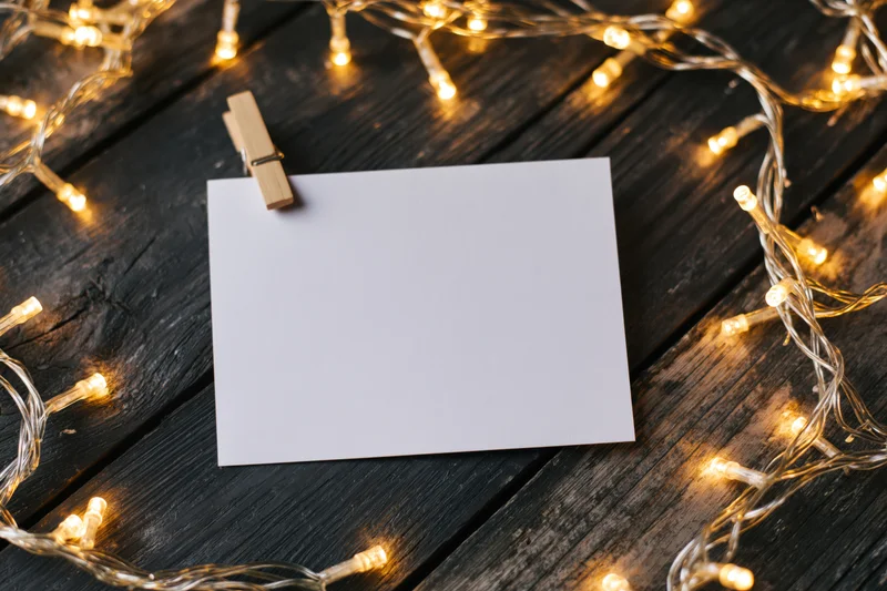 Blank white card clipped with wooden clothespin on dark wood surface surrounded by warm string lights