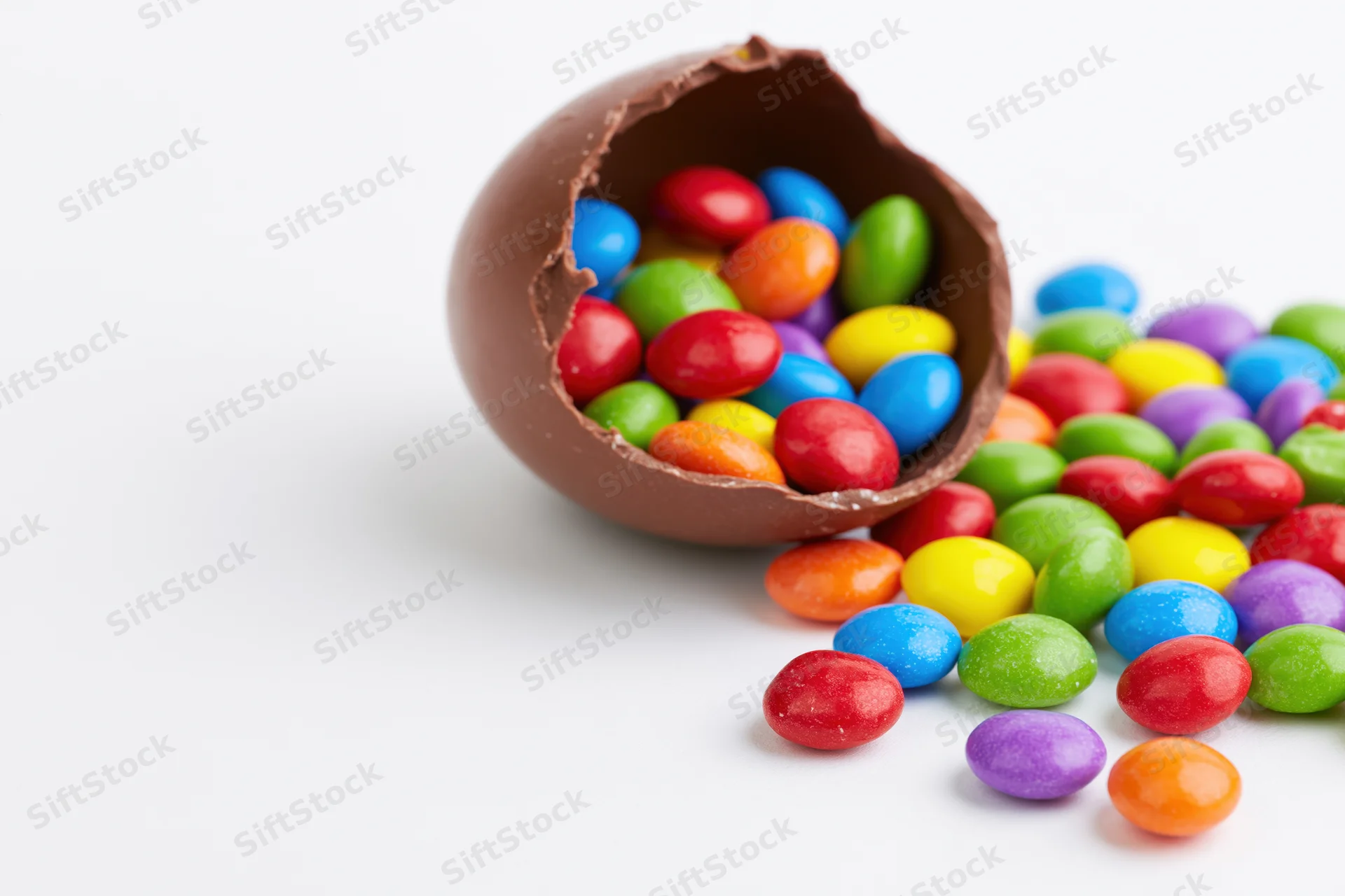 Broken chocolate easter egg spilling colorful candies onto white surface