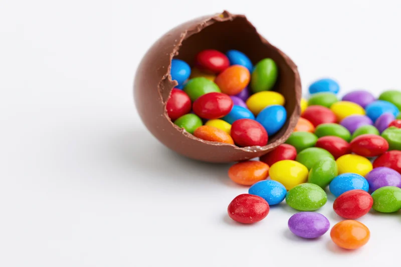 Broken chocolate easter egg spilling colorful candies onto white surface