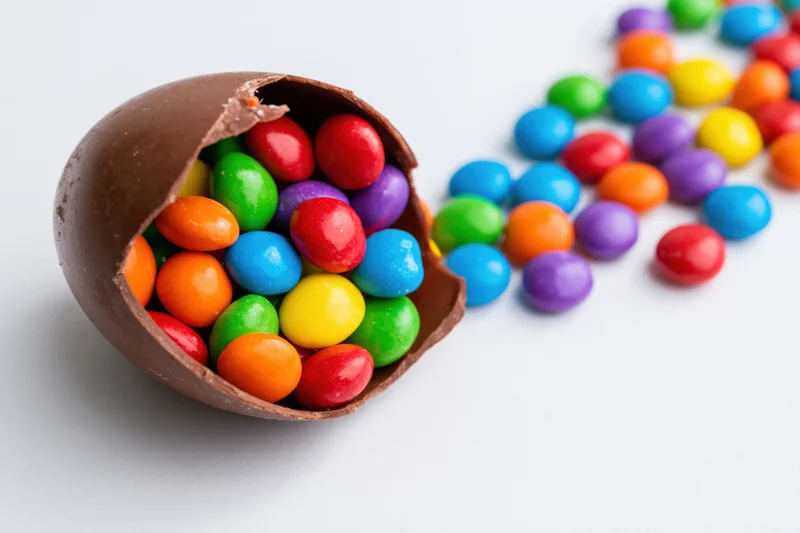 Broken chocolate easter egg shell spilling colorful candy onto white surface