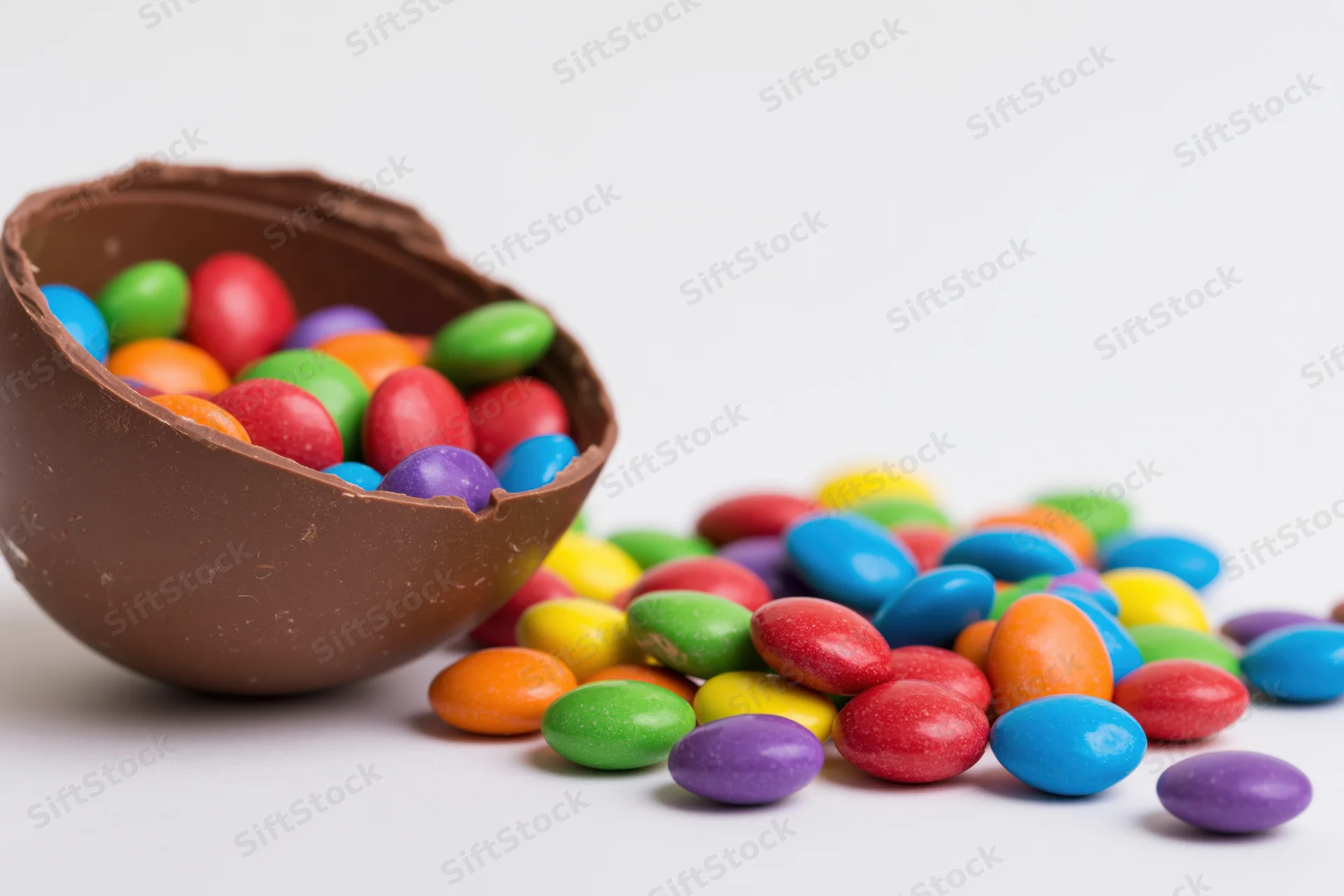 Broken chocolate easter egg shell spilling colorful candycoated sweets onto white surface