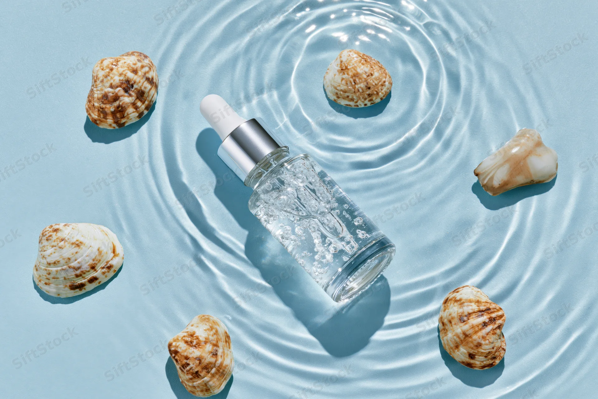 Clear glass dropper bottle with silver cap and seashells on rippled blue surface