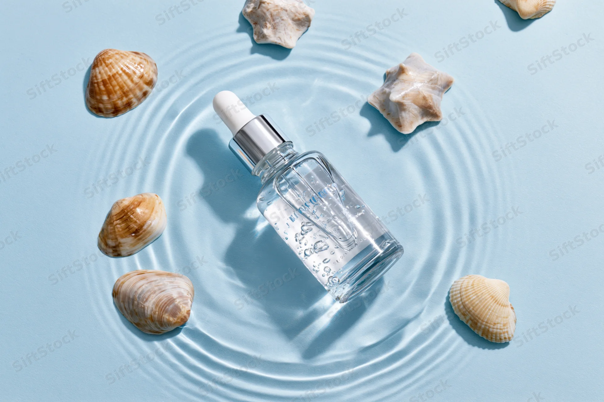 Clear glass dropper bottle with silver cap partially submerged in water ripples surrounded by seashells on light blue surface