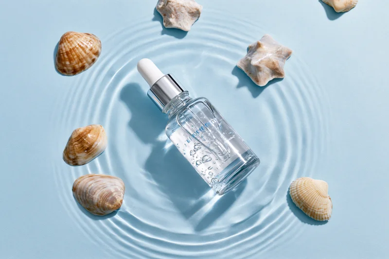 Clear glass dropper bottle with silver cap partially submerged in water ripples surrounded by seashells on light blue surface