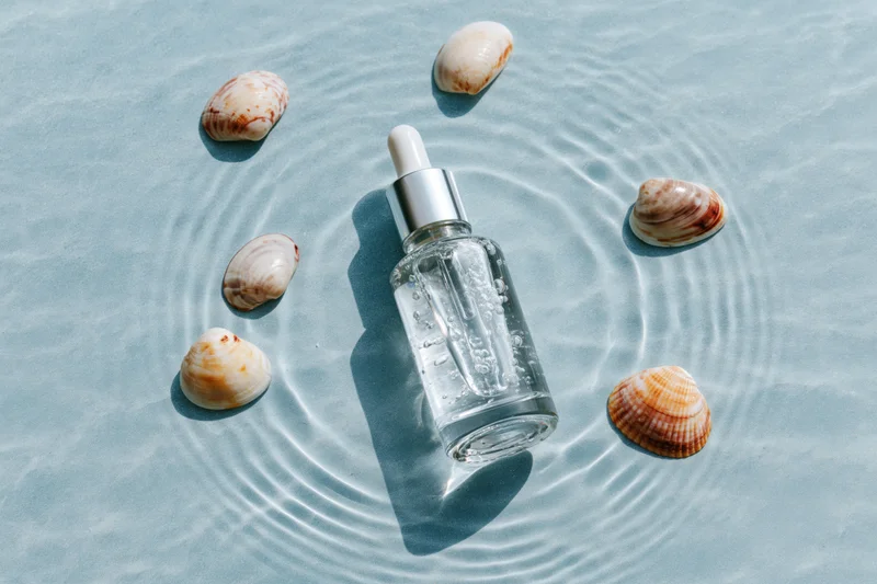Clear glass dropper bottle with silver cap surrounded by seashells in shallow water on sandy surface