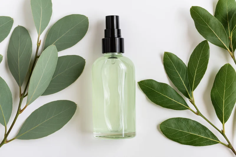 Clear glass spray bottle with pale green liquid and black pump surrounded by green leaves on white surface