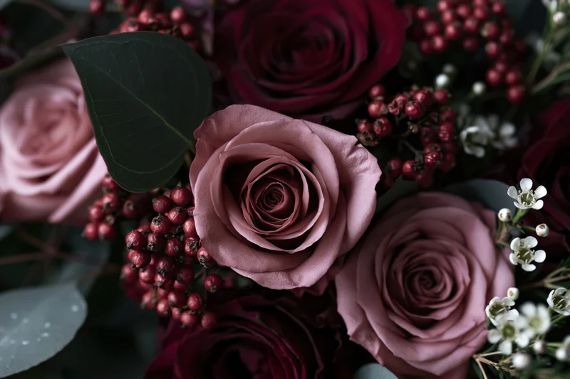 Close-up of a dense floral arrangement with dusty pink and deep red roses, small white flowers, red berries, and green foliage