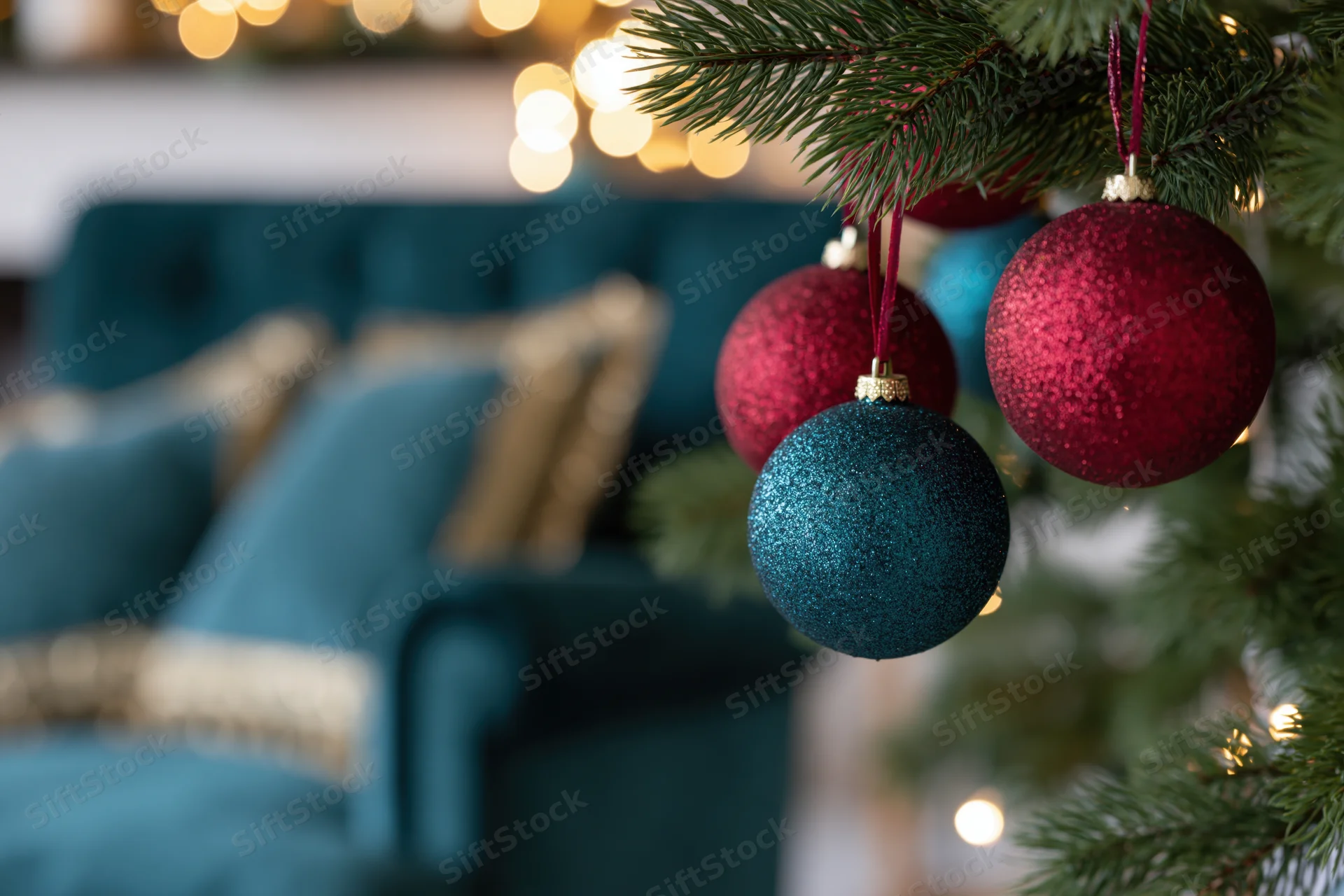 Close-up of glittery crimson and teal christmas ornaments hanging from a pine tree branch with blurred living room background