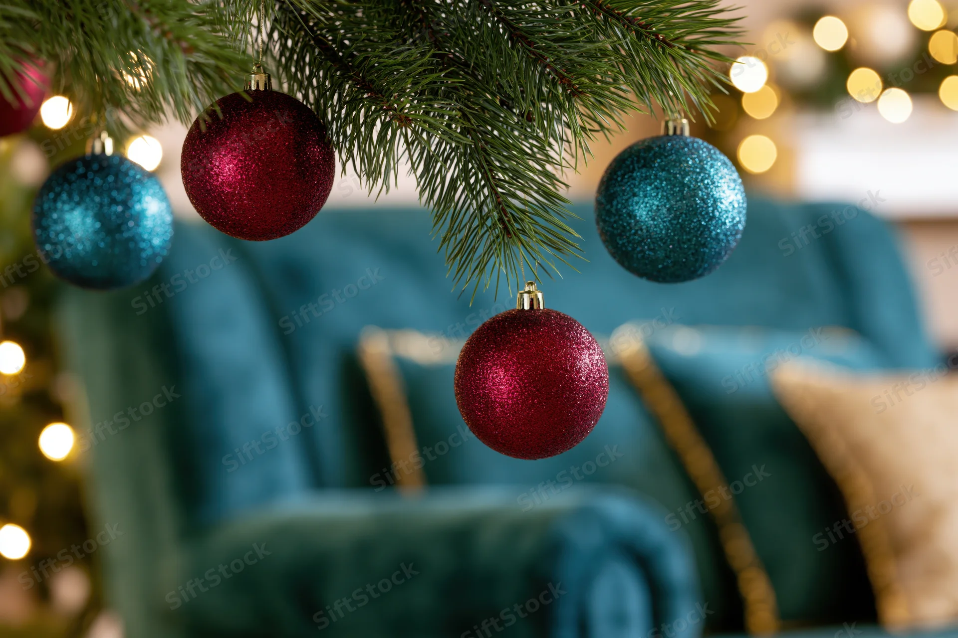 Close-up of glittery crimson and teal christmas ornaments hanging from a pine tree branch above a teal velvet sofa with gold-trimmed cushions and warm bokeh lights in the background
