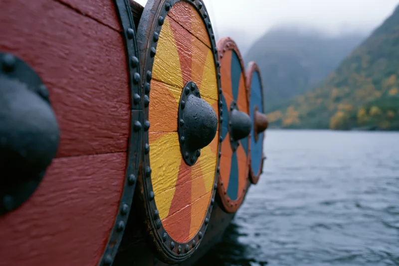 Viking longship hull with painted wooden shields lined along the side on a misty lake surrounded by autumn mountains