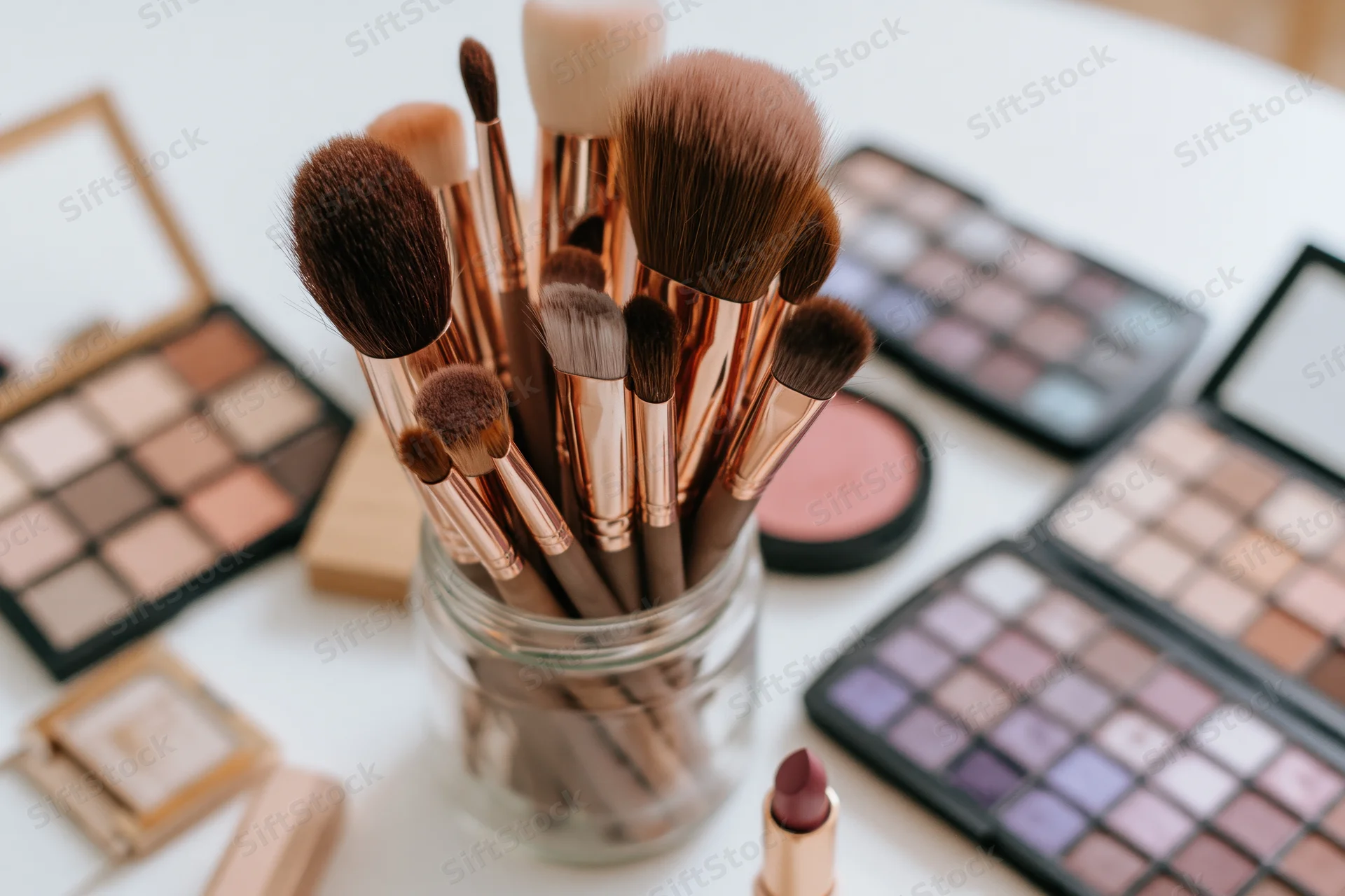 Makeup brushes in a glass jar surrounded by open palettes and cosmetics on a white surface