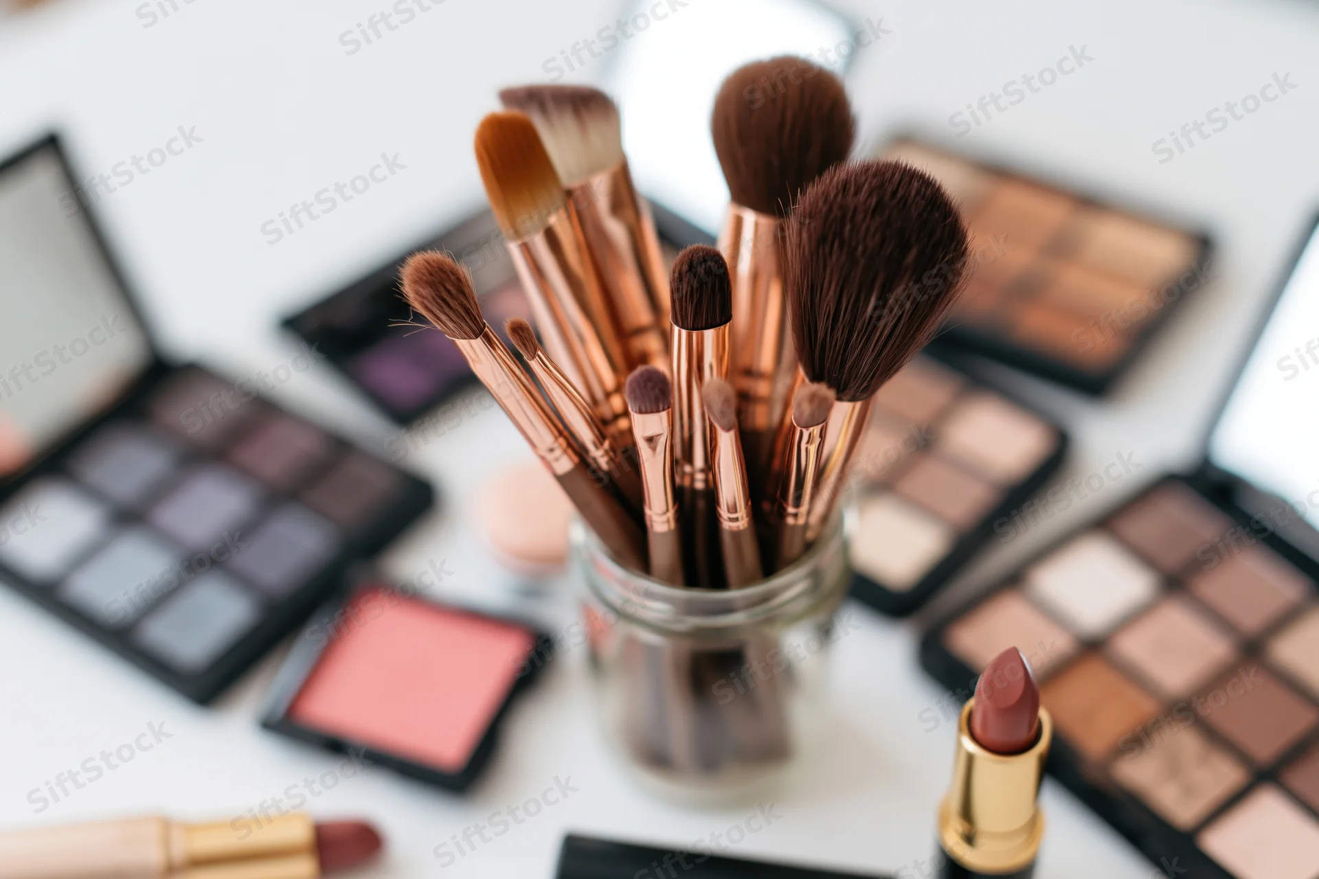 Makeup brushes in a glass jar surrounded by open palettes and lipsticks on a white surface