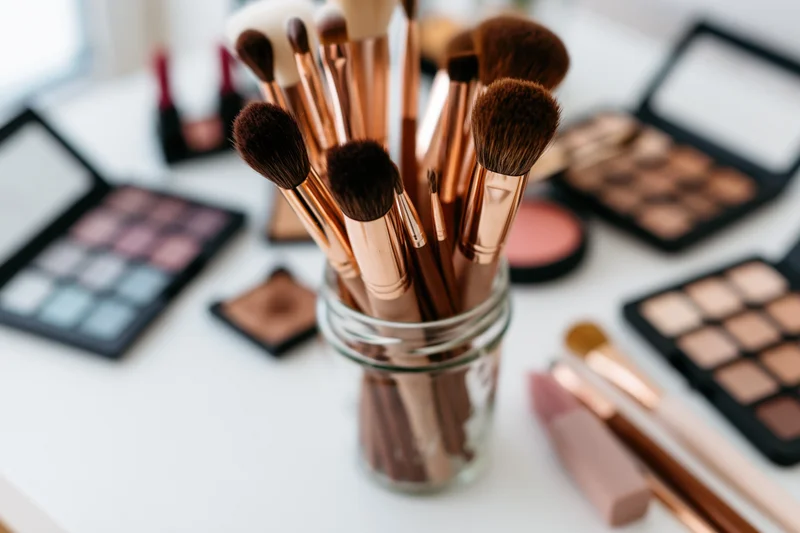 Makeup brushes in a glass jar surrounded by open palettes and cosmetics on a white table