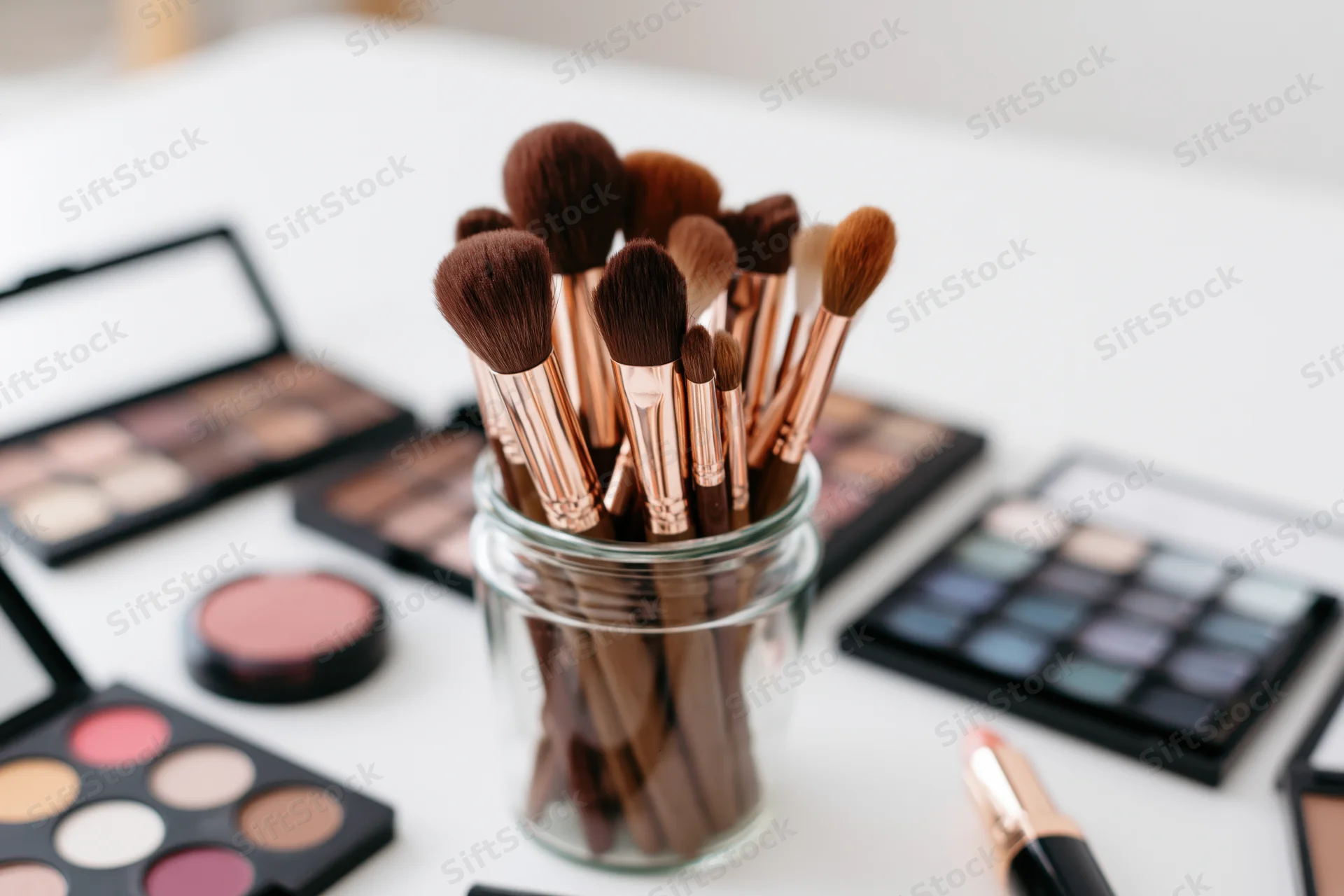 Makeup brushes in a glass jar surrounded by open palettes and cosmetics on a white table