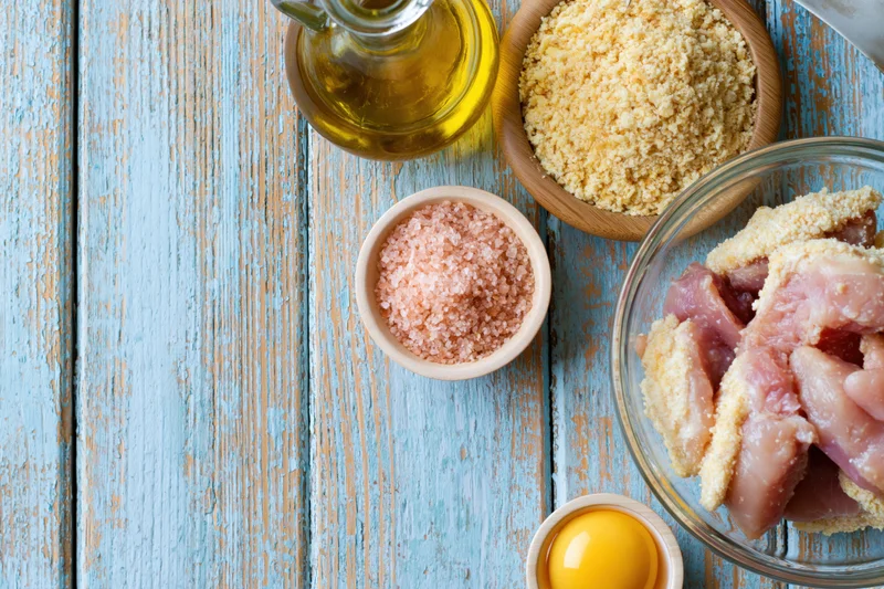 Raw chicken pieces breaded with egg, oil, salt and breadcrumbs arranged on a weathered blue wooden table in a flat lay composition